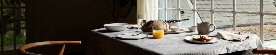 Morning dining table set with white plates, bread, orange juice and mug by a large window
