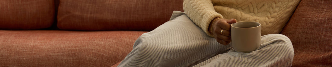 Person in cream knit sweater seated on terracotta sofa holding a matte beige ceramic mug