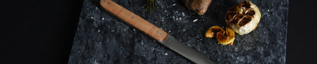 Steak knife with wood handle on dark marble board beside roasted garlic and herbs