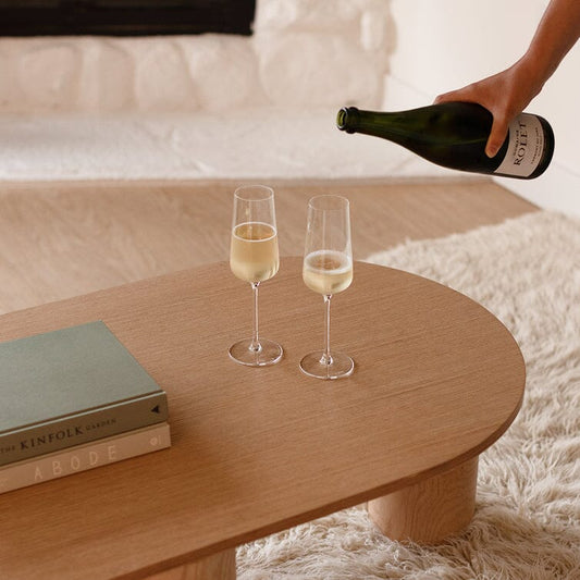Person pouring champagne into two glasses on a wooden coffee table with books and a bottle. #clear