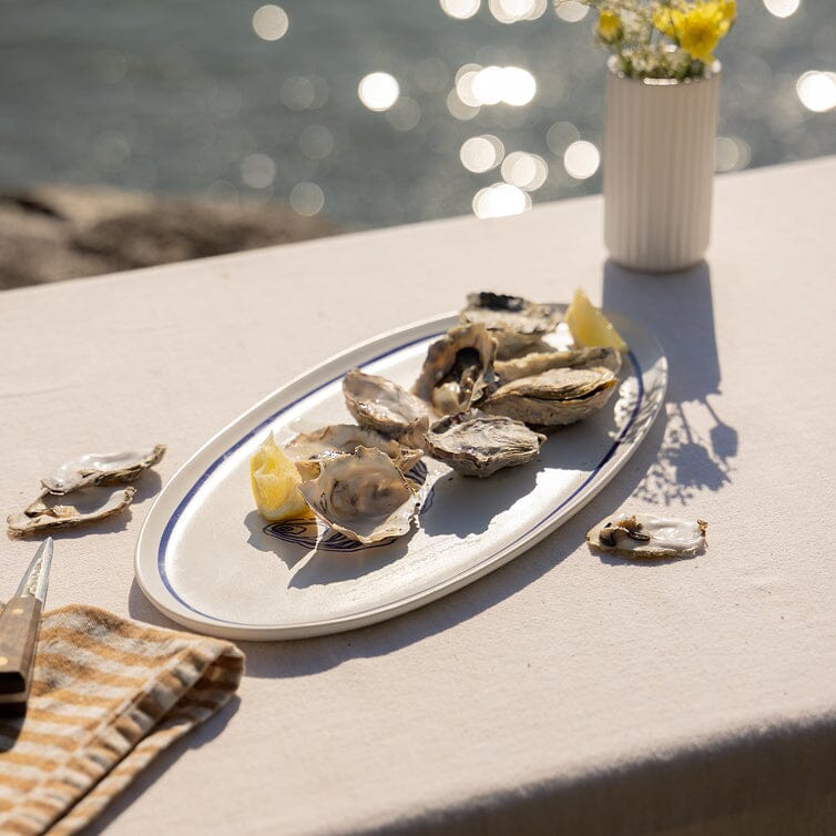 Platter of oysters on a table by a body of water with a vase of flowers.
